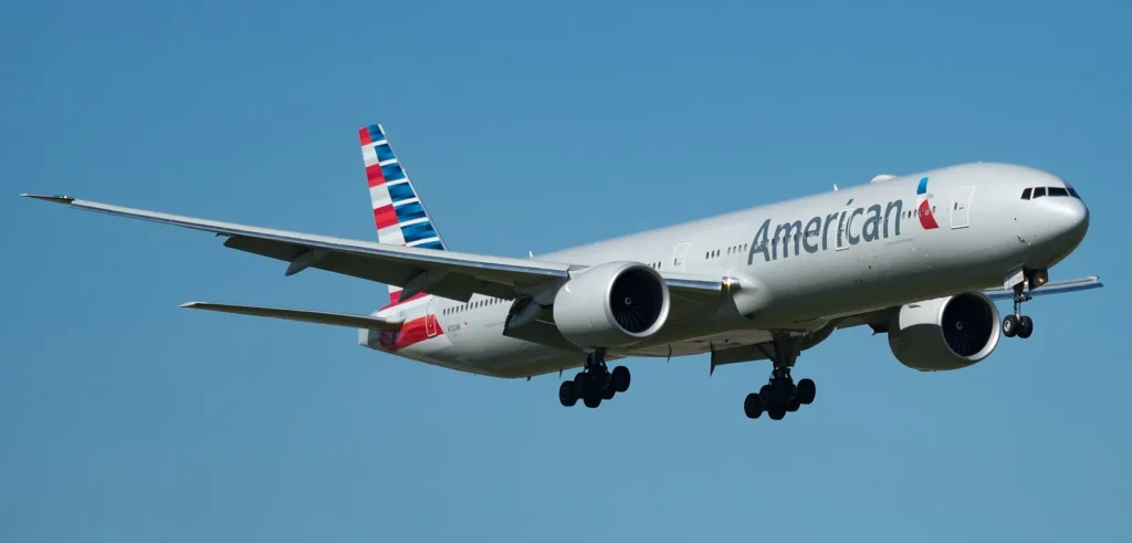 An American Airlines Boeing 777-300ER on final approach against a clear blue sky, landing gear extended.