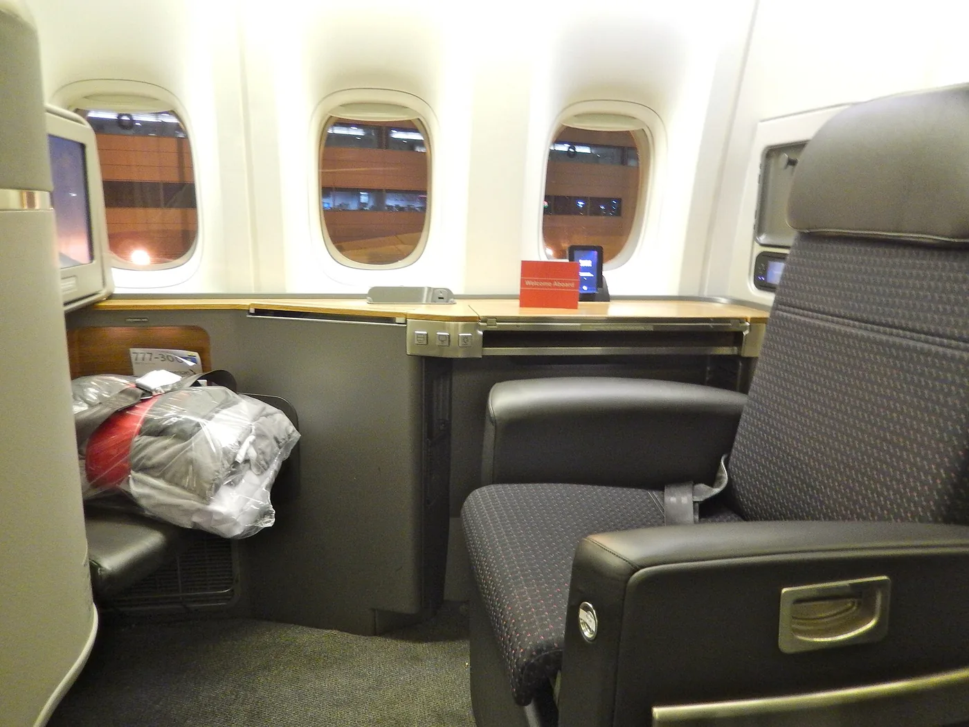 An American Airlines first class seat on a Boeing 777-300ER at night, with a wide side console, a folded bedding pack and a card reading Welcome Aboard.