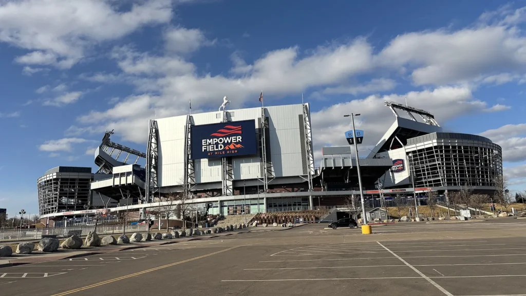 Exterior of Empower Field at Mile High in Denver on a clear day, with the stadium's white facade, a large Empower Field at Mile High sign and the bucking horse statue on top, seen across an empty parking lot.