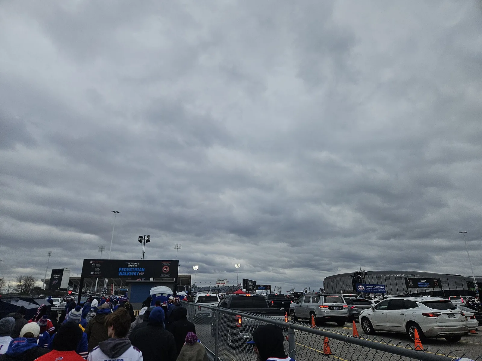 Bills fans in winter hats walk along a pedestrian walkway under a heavy gray sky at Highmark Stadium in Orchard Park, with the old stadium at left and the curved exterior of the new stadium beyond the parking lot at right