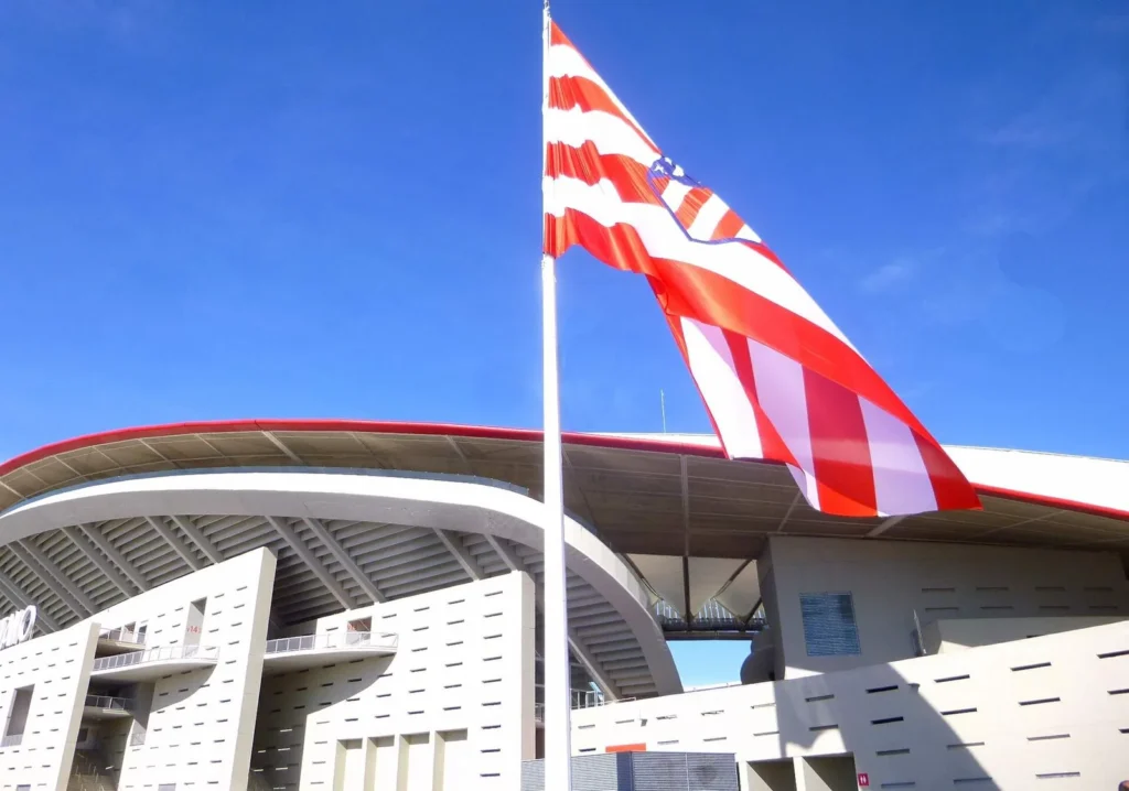 A giant red-and-white striped Atlético de Madrid flag flying on a tall white pole in front of the curved white facade and red roof edge of the Metropolitano stadium under a clear blue sky