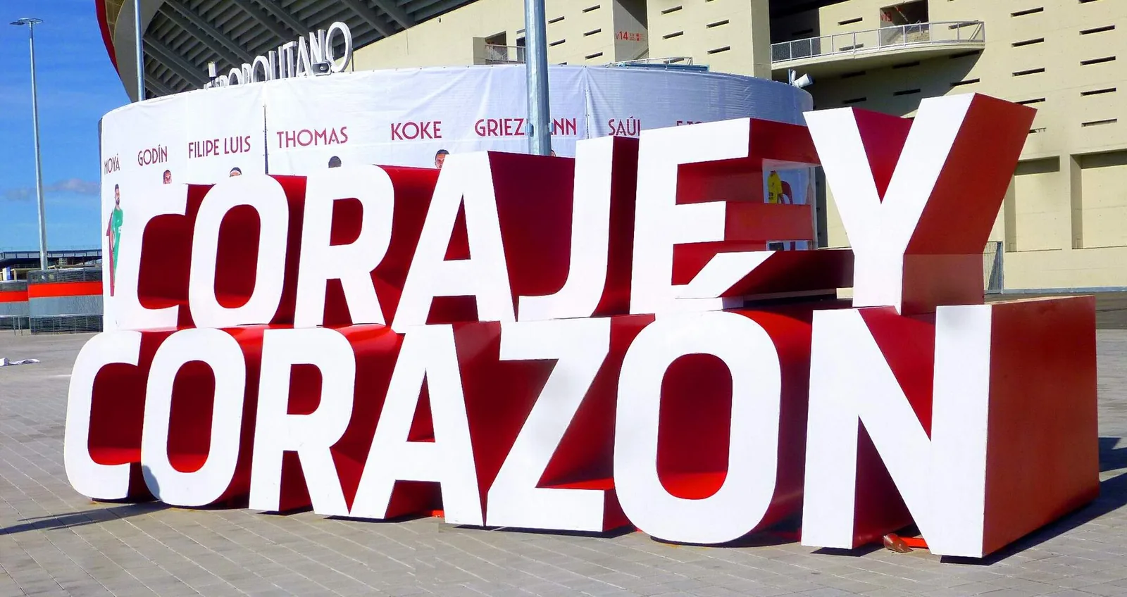 Large red and white 3D letters spelling "Coraje y Corazón" on the paved plaza outside the Metropolitano stadium, with a banner of Atlético players behind them