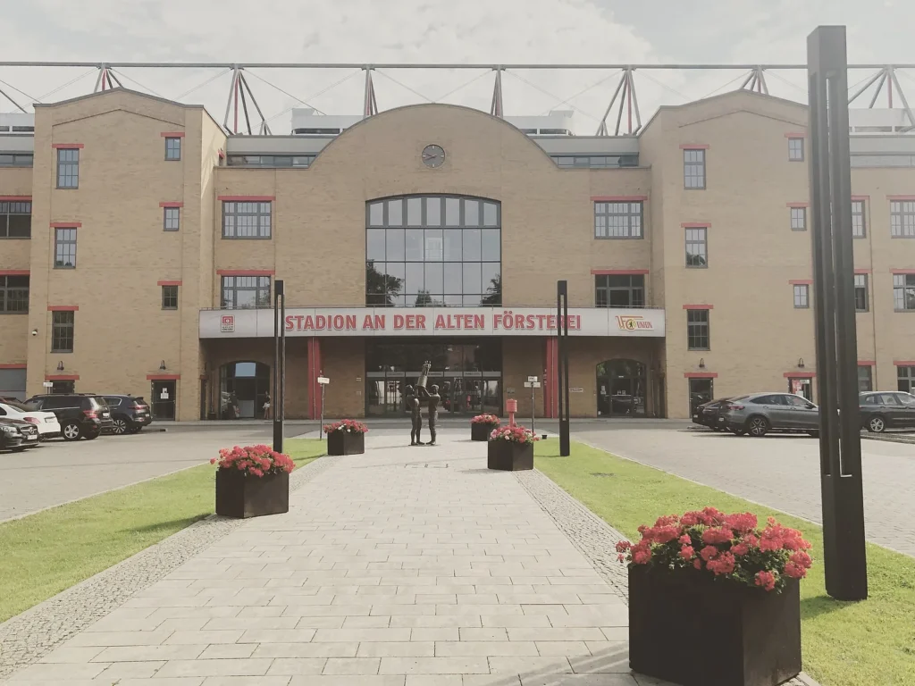 Brick main entrance of Stadion An der Alten Försterei in Berlin with the red stadium name sign above the doors, a paved approach and planters of red geraniums in front