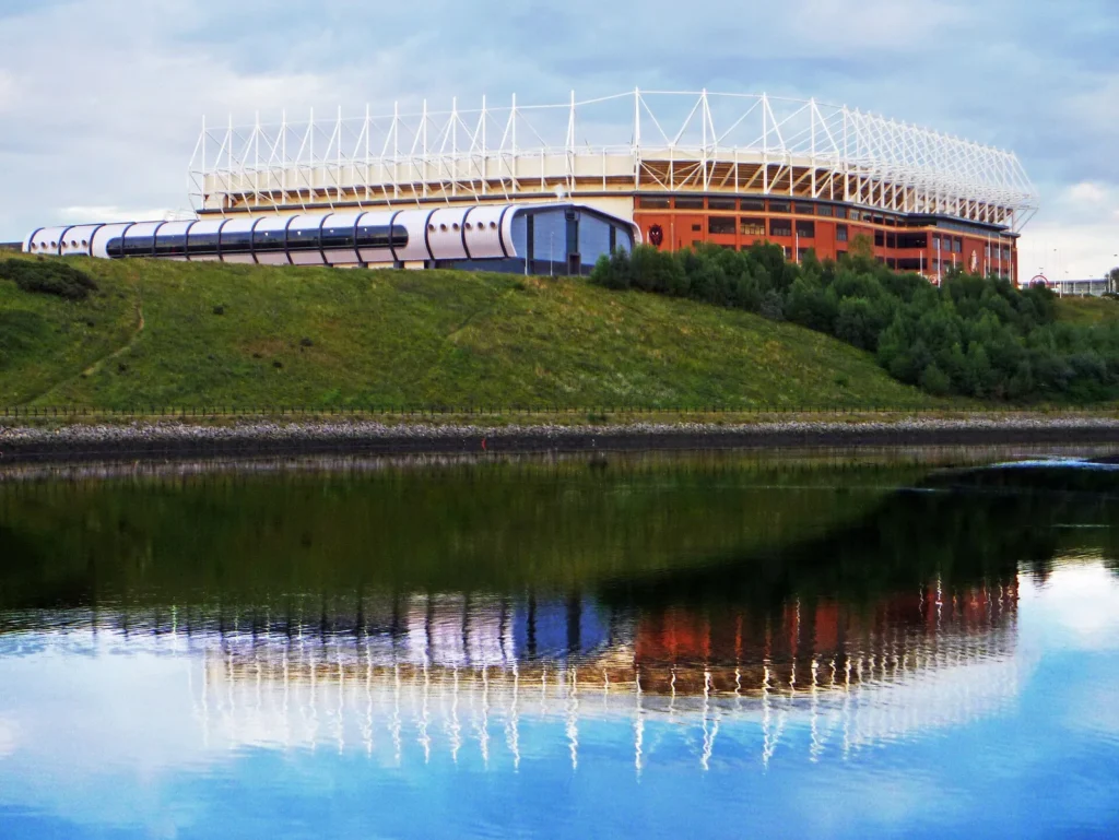 The Stadium of Light in Sunderland seen from across the River Wear, its white roof trusses and red-brick stand sitting on a grassy bank above the water, with a curved silver glass-and-metal building in front of it and the whole stadium mirrored upside down in the calm river under a cloudy blue sky.
