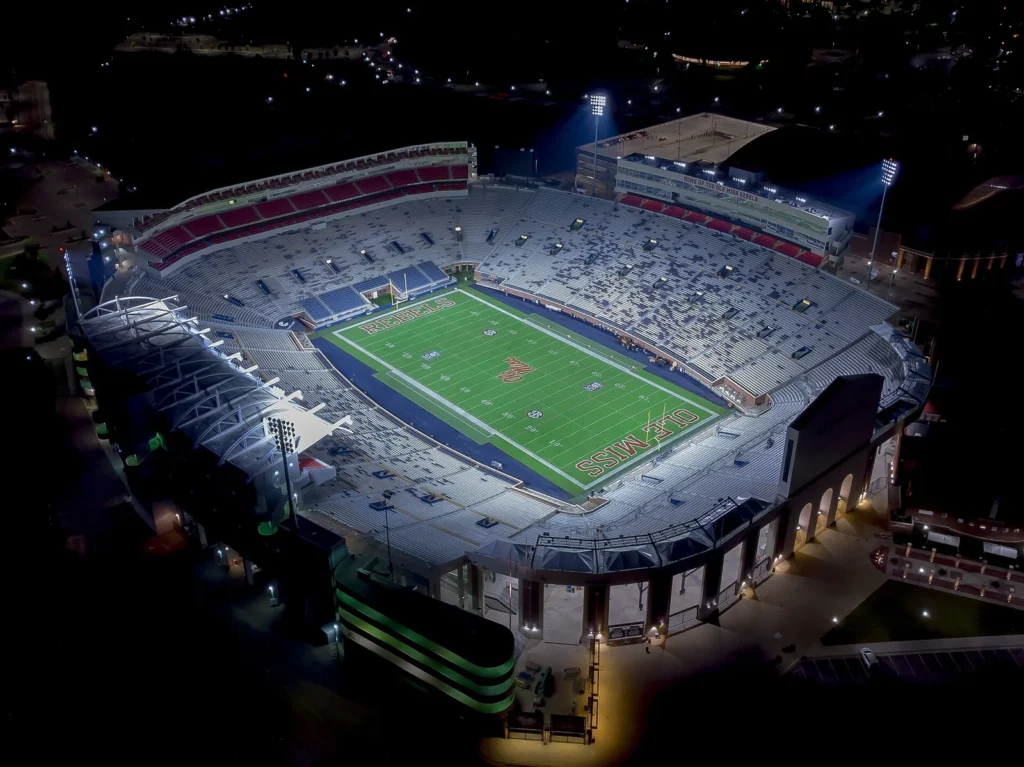 Aerial night view of Vaught-Hemingway Stadium in Oxford, Mississippi, with the green field marked REBELS and OLE MISS in the end zones, lit stands and the surrounding campus in the dark