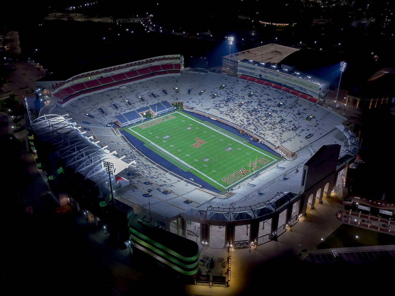 Aerial night view of Vaught-Hemingway Stadium in Oxford, Mississippi, with the green field marked REBELS and OLE MISS in the end zones, lit stands and the surrounding campus in the dark