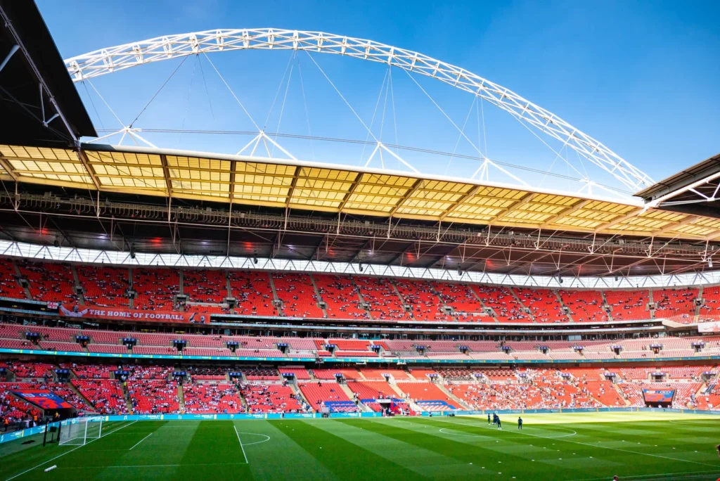Inside Wembley Stadium on a sunny day, with tiers of red seats around the pitch and the stadium's white arch curving over the roof