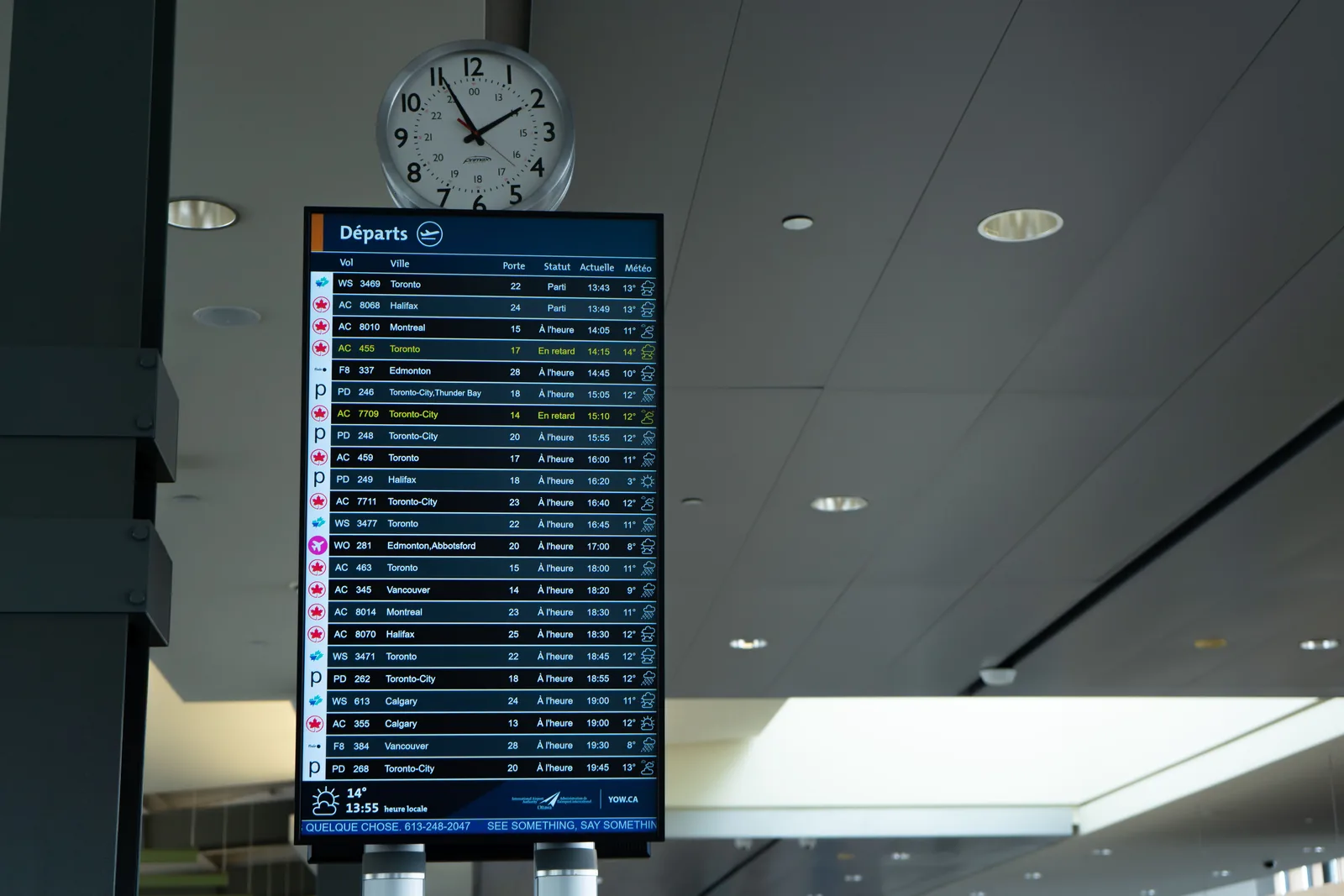 An airport departure board listing flight times and gates, mounted below a large round analogue wall clock.