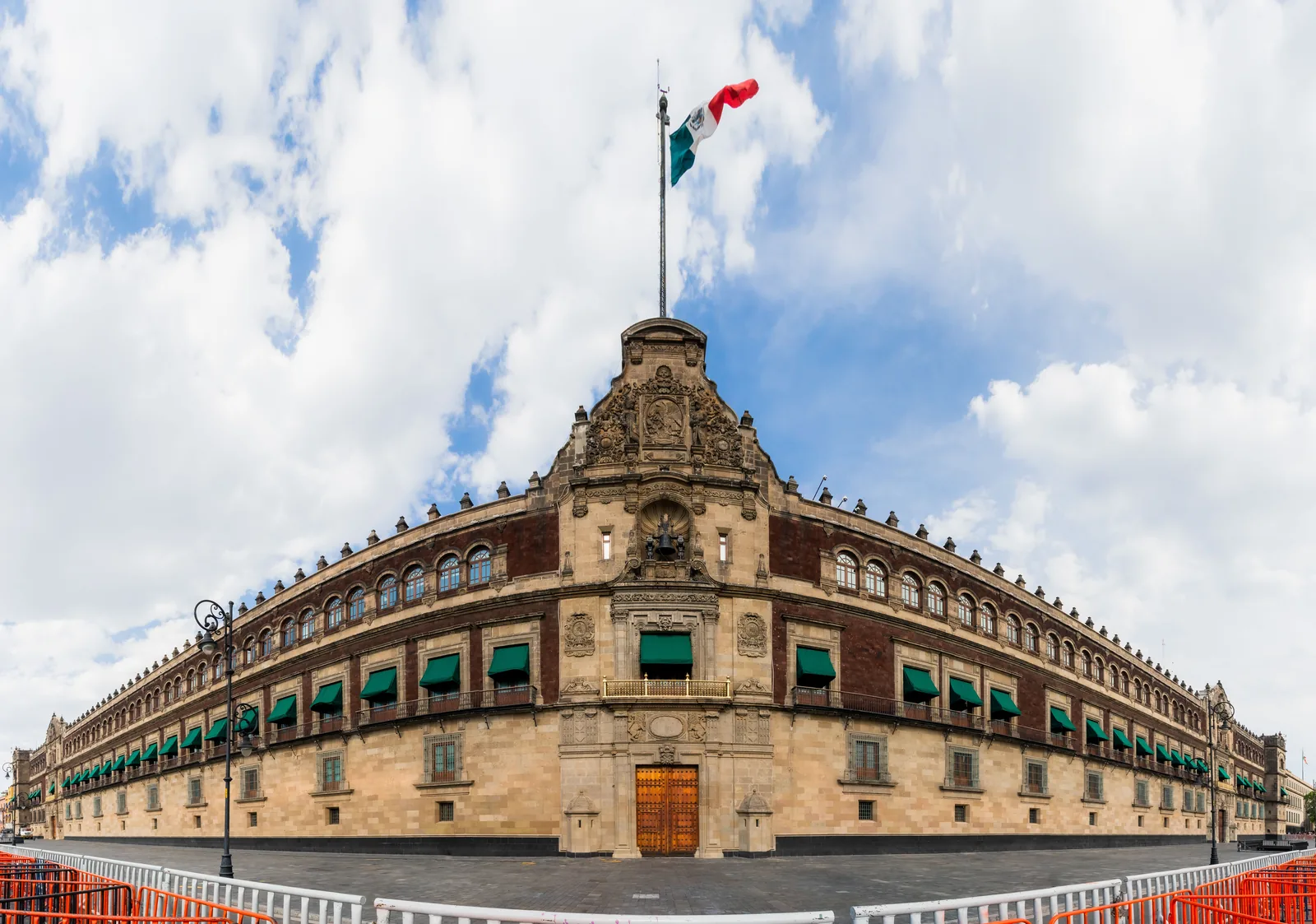 The stone and red tezontle facade of the National Palace in Mexico City under a cloudy blue sky, with a large Mexican flag flying above the central tower, the bell set in a niche over the gilded central balcony, rows of green window awnings along both wings and crowd barriers in the foreground.