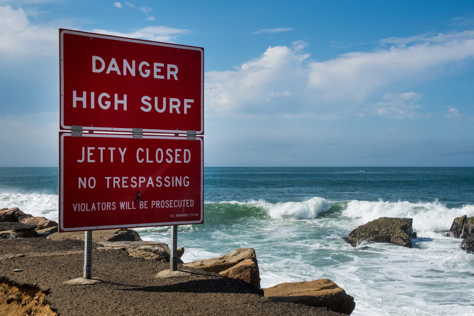 A red warning sign reading DANGER HIGH SURF above a second panel reading JETTY CLOSED, NO TRESPASSING, standing on a rocky breakwater while waves break against the boulders behind it under a blue sky.