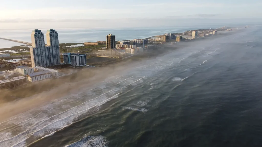 Aerial view of South Padre Island, Texas, with high-rise hotels along the beach, waves rolling onto the sand and a low sea mist drifting over the shoreline.
