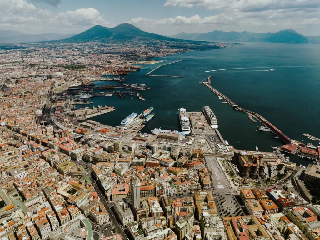 Aerial view of Naples: dense terracotta rooftops, the harbour with ferries and cruise ships at the quays, and the twin summit of Mount Vesuvius rising across the bay under scattered cloud.
