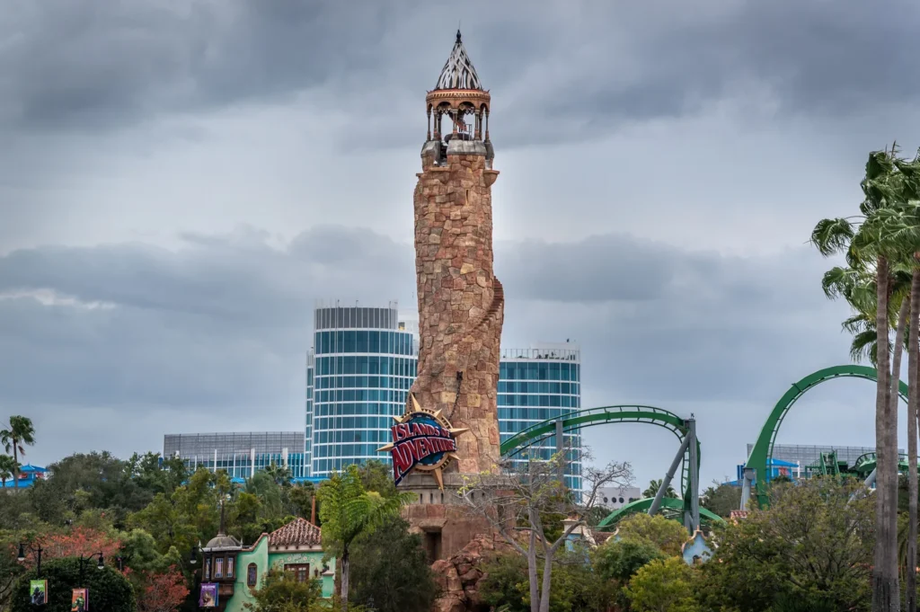 The stone lighthouse tower with the Islands of Adventure sign at the entrance to the Universal Orlando park, with the green track of a roller coaster, palm trees and tall glass buildings behind under grey clouds