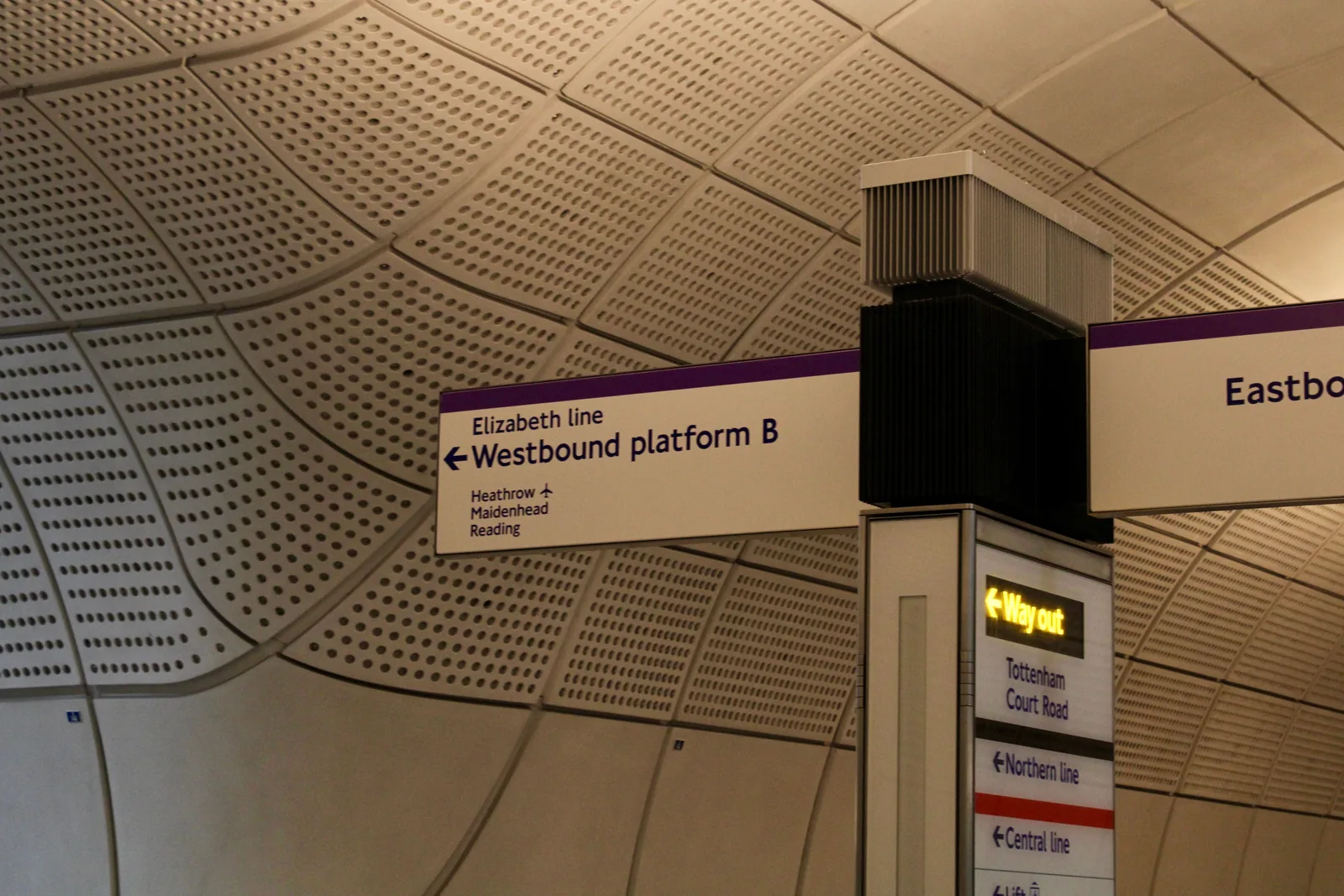 Elizabeth line signage under the curved, perforated concrete ceiling of a London station: a white sign with a purple band points left to the westbound platform for Heathrow, Maidenhead and Reading, with an eastbound sign and a yellow Way out sign beyond it.