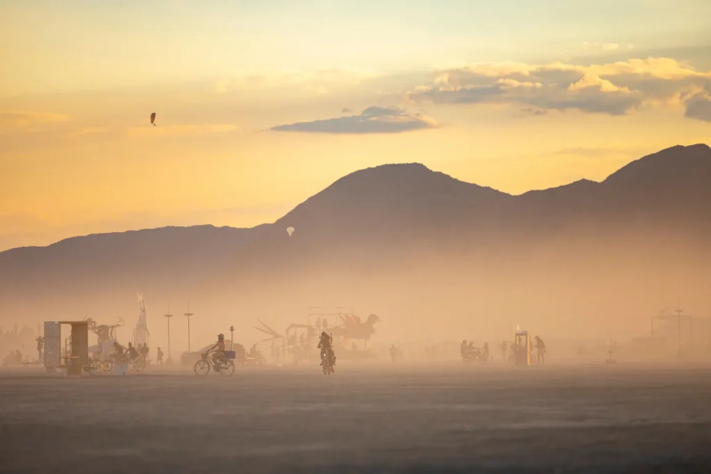 Cyclists, art cars and installations spread across the open desert playa at sunset, seen through dust haze with mountains and a hot-air balloon on the horizon.