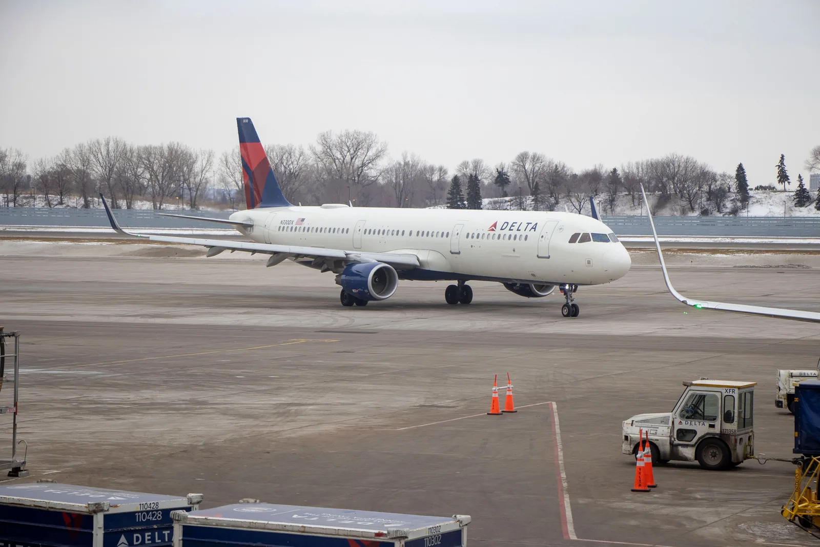 A Delta Air Lines narrow-body jet taxis across a snow-edged airport apron on a gray winter day, with orange cones and a Delta ground vehicle in the foreground.