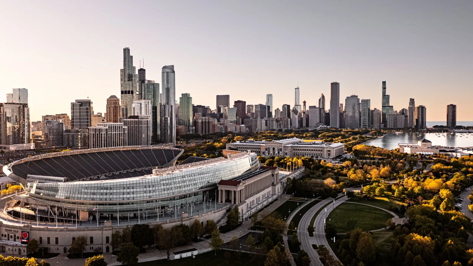 Aerial view of Soldier Field at golden hour, its glass-and-steel bowl rising above the original stone colonnade, with the Field Museum behind it, the Shedd Aquarium beside the harbor on the right and the Chicago skyline beyond trees turning yellow.
