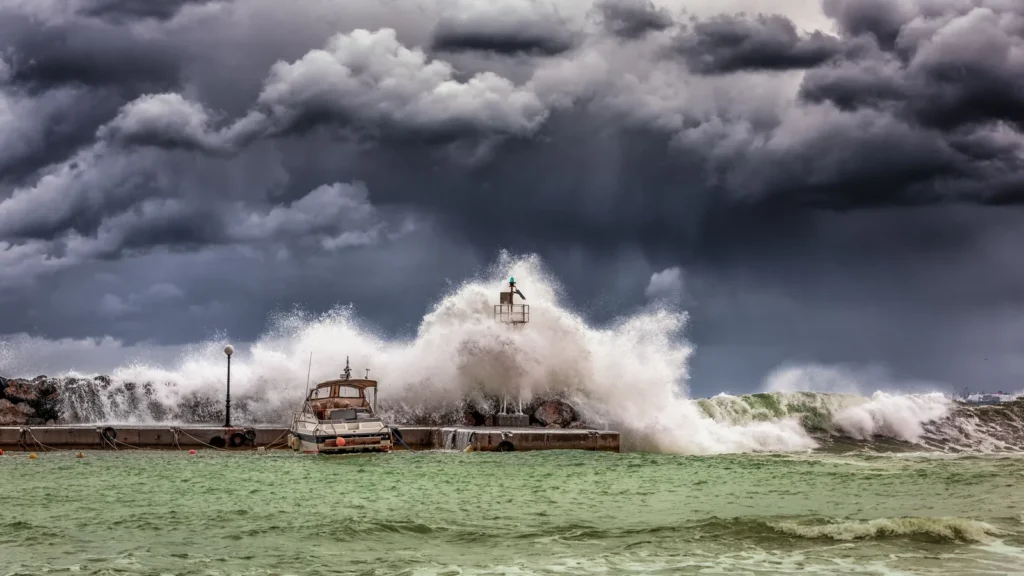 A large wave breaks over a harbour breakwater beside a small moored boat, under heavy grey storm clouds.