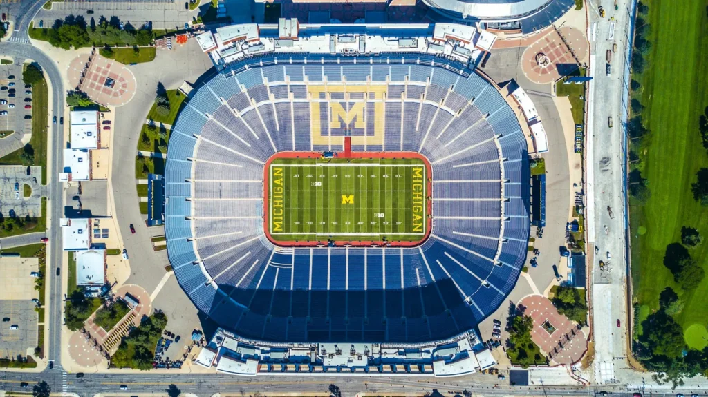Top-down aerial view of Michigan Stadium in Ann Arbor on a sunny day with the stands empty: a bowl of blue bleacher seats around a green football field with MICHIGAN painted in maize letters in both end zones, a large block M picked out in the seats, and the round roof of Crisler Center at the top edge.
