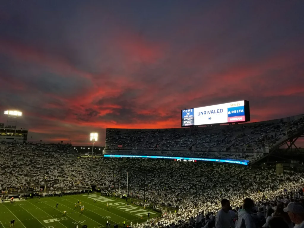 Beaver Stadium packed with fans dressed in white during a Penn State night game, under a sunset sky streaked with red clouds, with the lit video board above the stands and the field below.