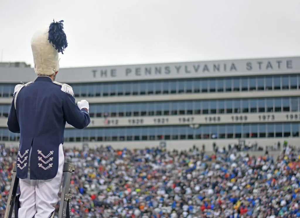 A drum major in a navy uniform and tall white plumed hat stands on a ladder facing a crowded Beaver Stadium grandstand, below a facade lettered The Pennsylvania State and a row of years.