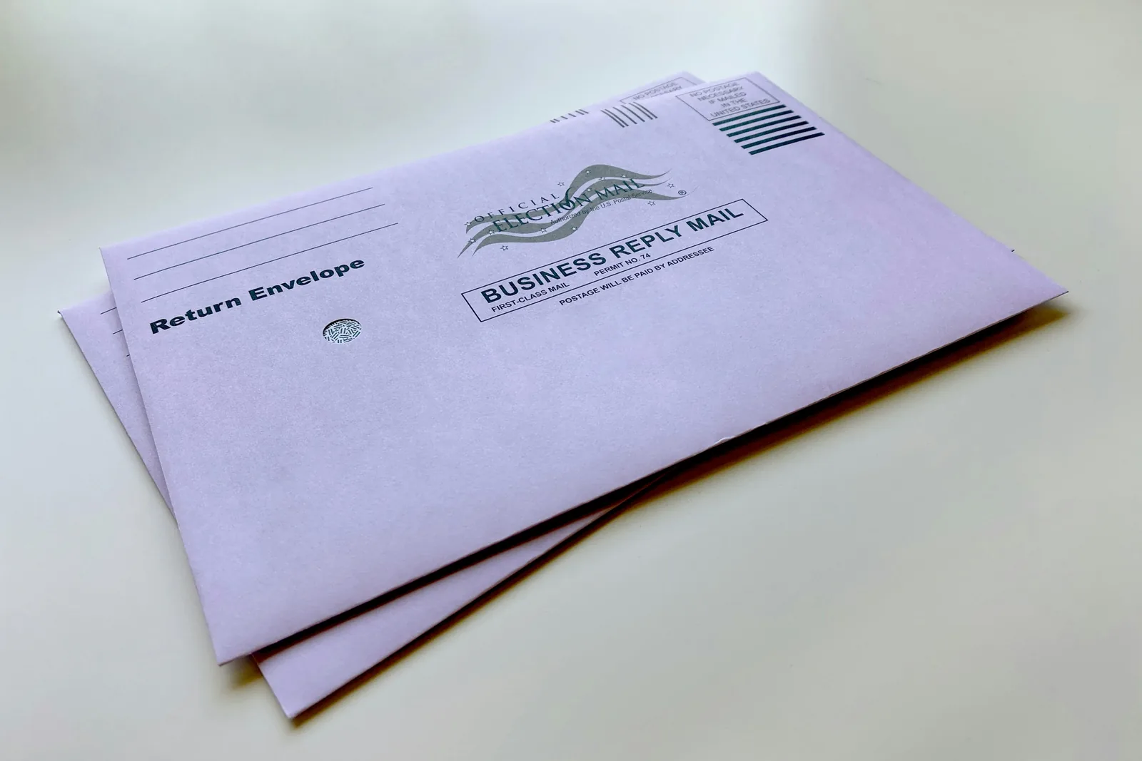Two pale purple absentee ballot return envelopes marked Official Election Mail and Business Reply Mail lie on a plain white surface.