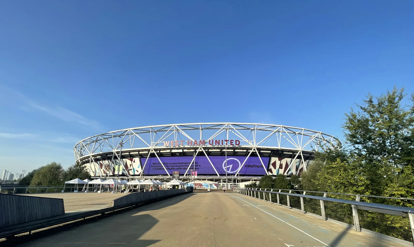 The London Stadium seen head-on from a raised pedestrian walkway in the Queen Elizabeth Olympic Park, its white lattice roof truss carrying the words WEST HAM UNITED above a purple hoarding, with the towers of Canary Wharf small on the horizon to the left.