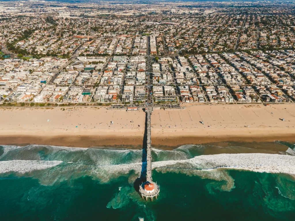 Aerial view of a long pier with a small red-roofed building at its tip, reaching out from a wide sandy beach into turquoise surf, with a dense grid of homes stretching inland behind it.