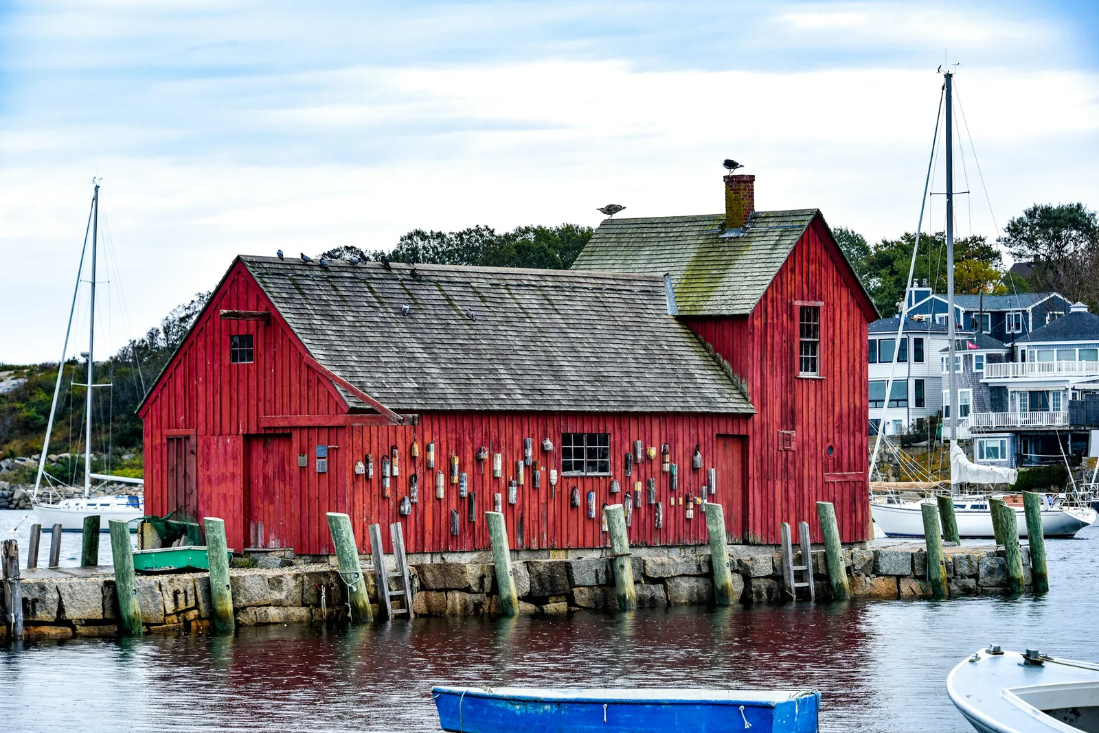 The red clapboard fish house known as Motif No. 1 stands on a granite pier in Rockport Harbor, its wall hung with lobster buoys, with moored sailboats and shingled houses behind it.