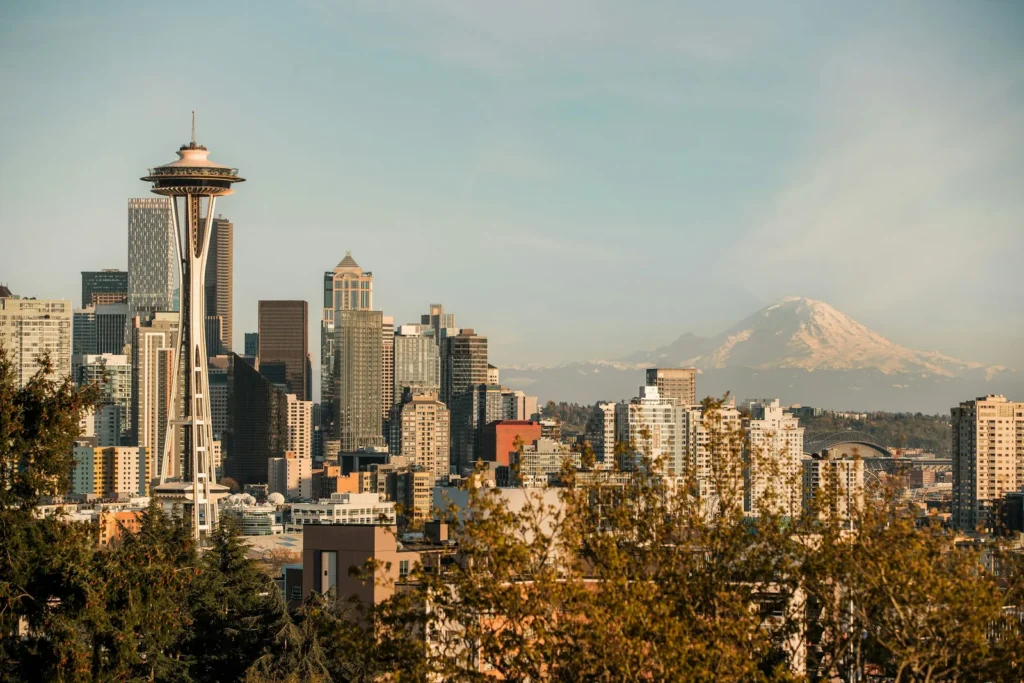 The Seattle skyline seen from Kerry Park in warm evening light: the Space Needle on the left in front of downtown towers, the snow-covered cone of Mount Rainier on the horizon to the right, and the roof of a stadium visible beyond the buildings.