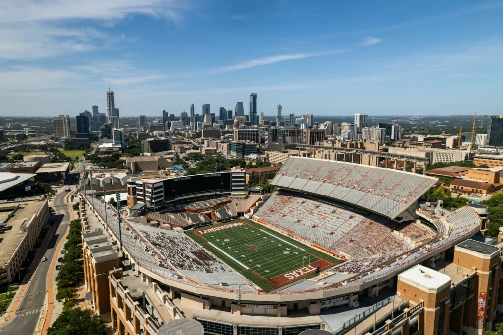 Aerial view of the empty Darrell K Royal-Texas Memorial Stadium on the University of Texas campus on a sunny day, with LONGHORNS and TEXAS painted in burnt orange end zones and a longhorn logo at midfield, stands enclosing the field with a large video board at one end, and downtown Austin's skyline with the dome of the Texas State Capitol beyond.