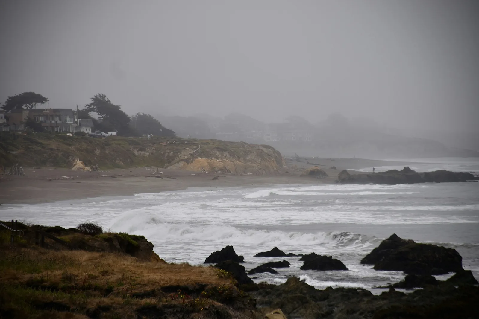 A grey, fog-bound stretch of the California coast at Cambria: lines of white surf breaking over dark rocks in the foreground, a low eroded bluff behind the beach, and houses among wind-bent cypress trees fading into the mist.