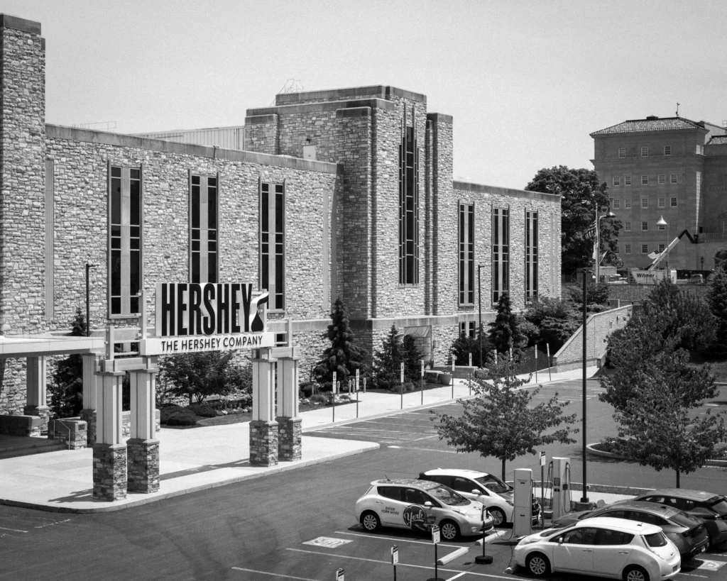 Black-and-white photo of a large stone building with tall narrow windows and a Hershey, The Hershey Company sign over its entrance canopy, with cars parked at charging stations in the lot in front.