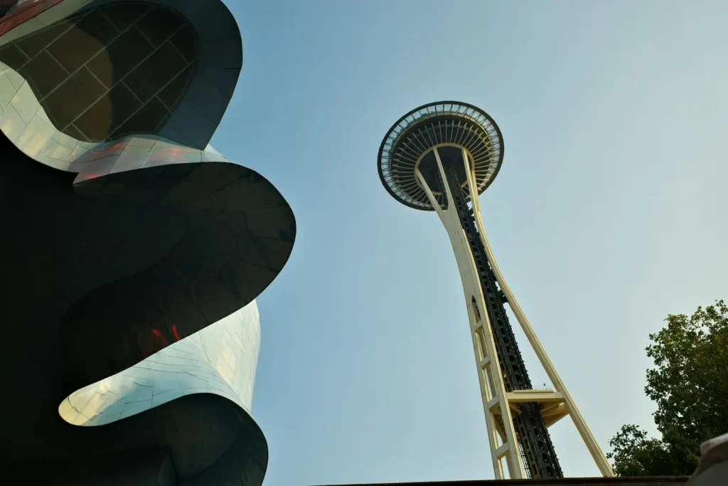 Looking up at the curved metallic facade of the Museum of Pop Culture in Seattle, with the Space Needle rising into a pale blue sky beside it.