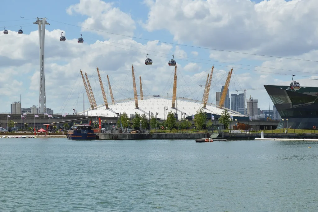 The O2 arena on the Greenwich Peninsula in London, its white roof and yellow masts seen across the water with cable cars passing overhead