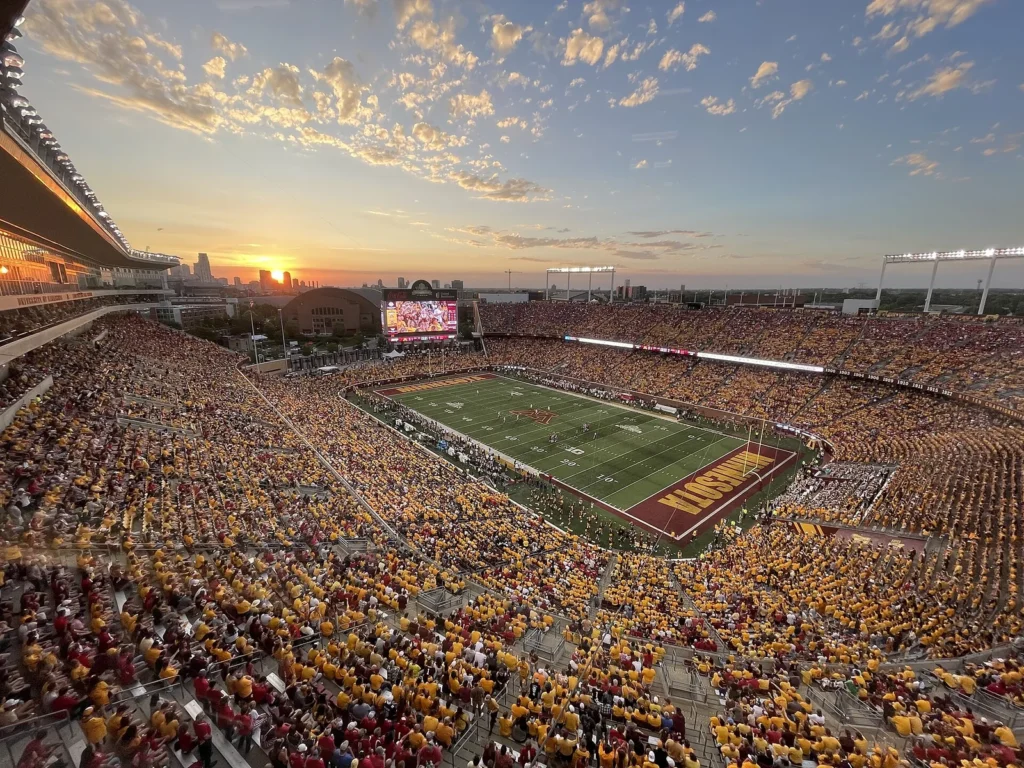Huntington Bank Stadium in Minneapolis at sunset, with fans in maroon and gold filling the stands and the city skyline glowing on the horizon