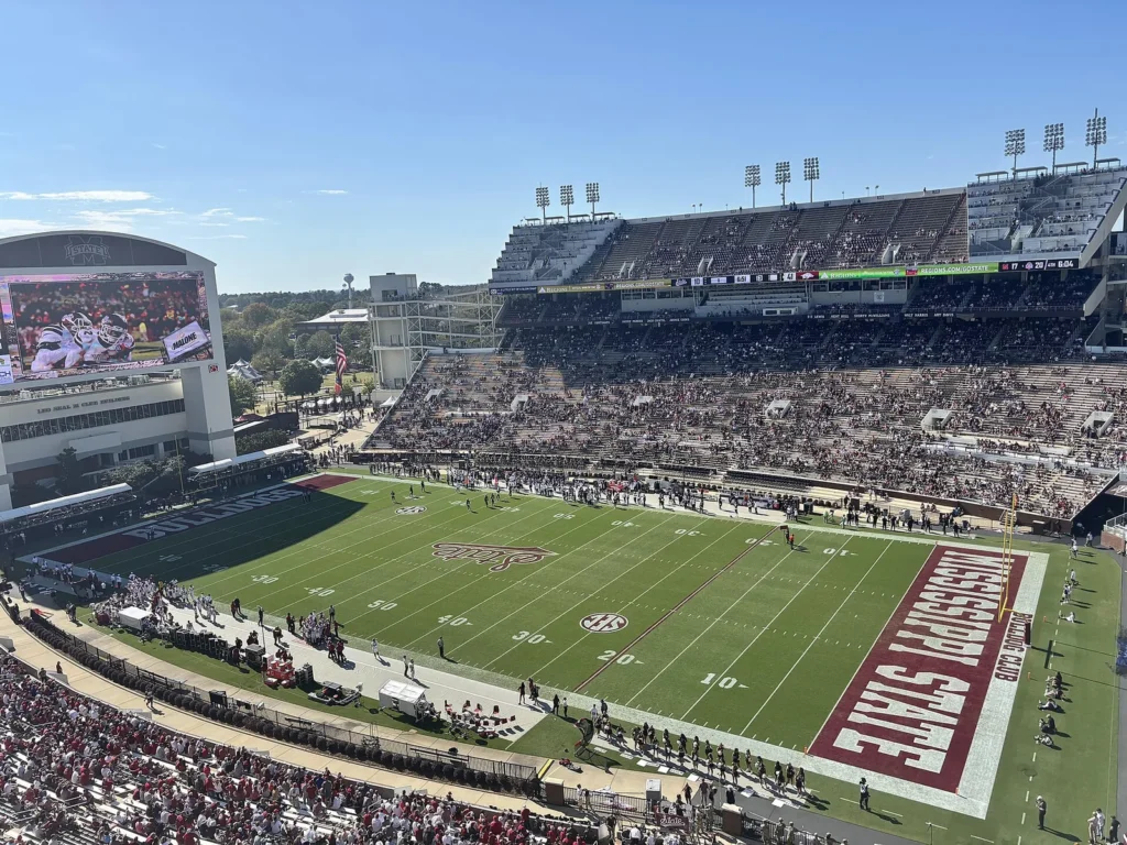 View from the upper deck of Davis Wade Stadium in Starkville, Mississippi, looking across the field toward a large videoboard and the packed east stands under a clear blue sky